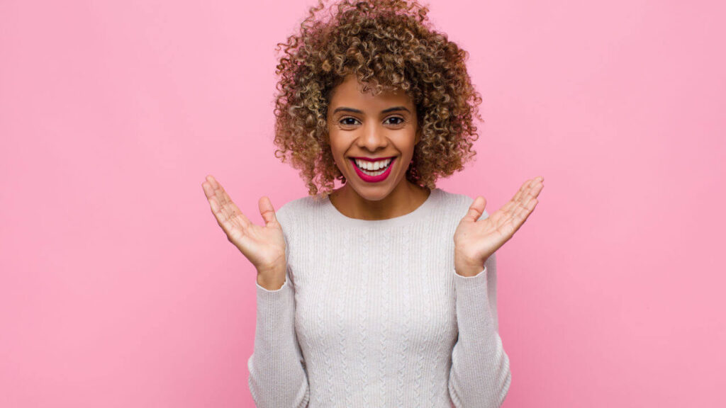 Jovem parecendo feliz e animada, com as duas mãos abertas ao lado do rosto, contra uma parede rosa.