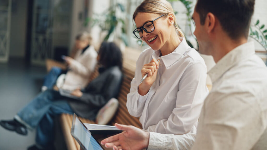 dupla de colegas de trabalho sentados olhando para a tela de um notebook e o fundo é um escritório desfocado