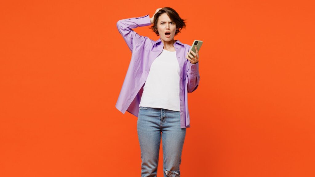 mulher no centro da imagem com a mão na cabeça e segurando um celular com a outra mão em fundo laranja