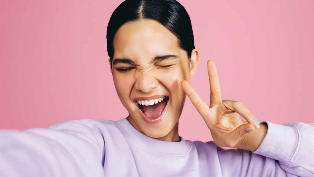 Jovem animada, tirando uma selfie em um estúdio, fazendo sinal de paz, com uma expressão de alegria no rosto. Mulher se divertindo, capturando seu momento de autoconfiança e positividade.