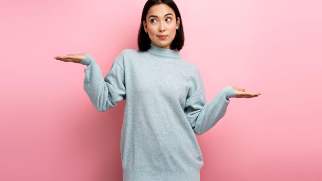 Retrato de mulher confusa e incerta, de suéter, expressando dúvidas, sem olhar para a câmera. Estúdio interno com fundo rosa.