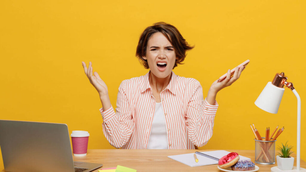 Jovem indignada, irritada, sentada na mesa de um escritório, segurando celular móvel, espalhando as mãos, isolada no fundo de cor amarela.
