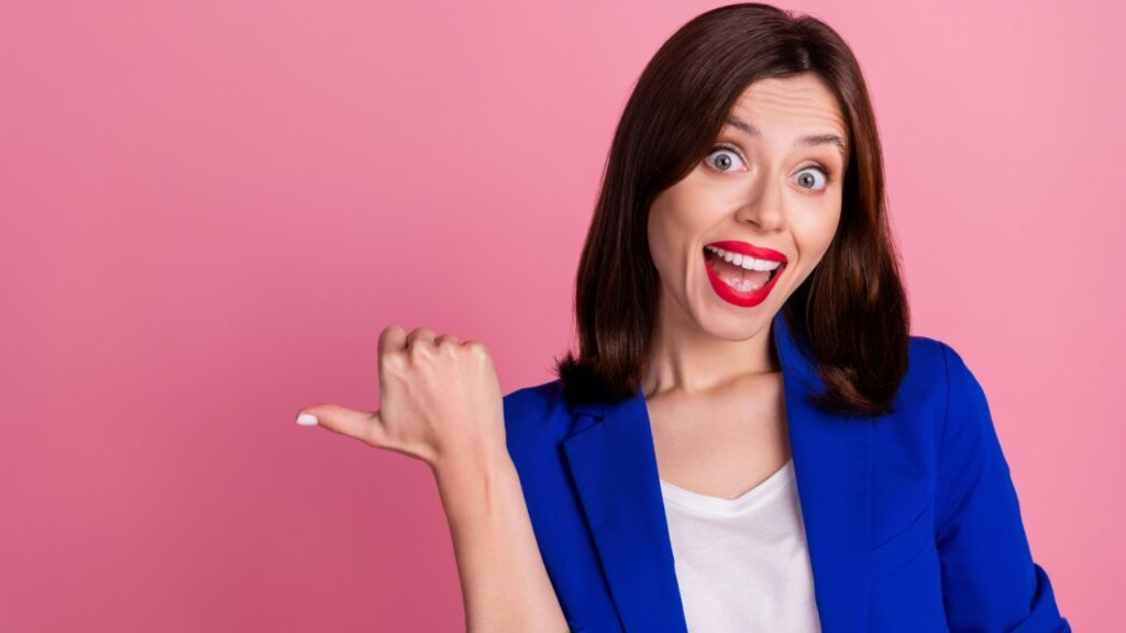 mulher sorrindo e apontando o polegar para o lado esquerdo em fundo rosa