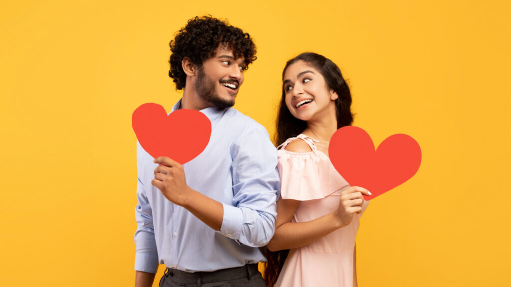 casal heterossexual de costas um para o outro e ambos sorrindo e segurando um coração vermelho em fundo amarelo