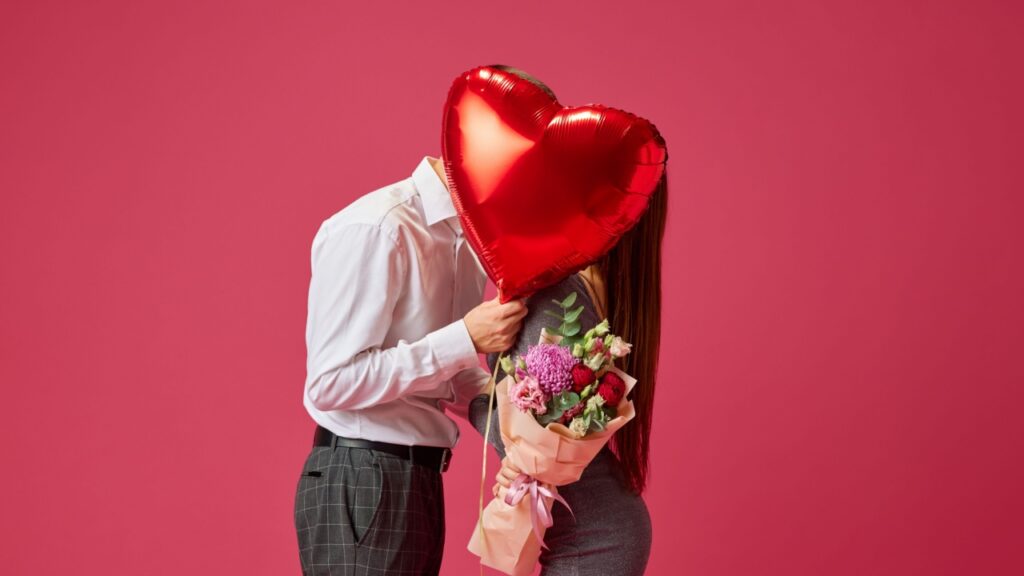 casal com roupas sociais tampando o rosto com um balão vermelho em fundo rosa queimado