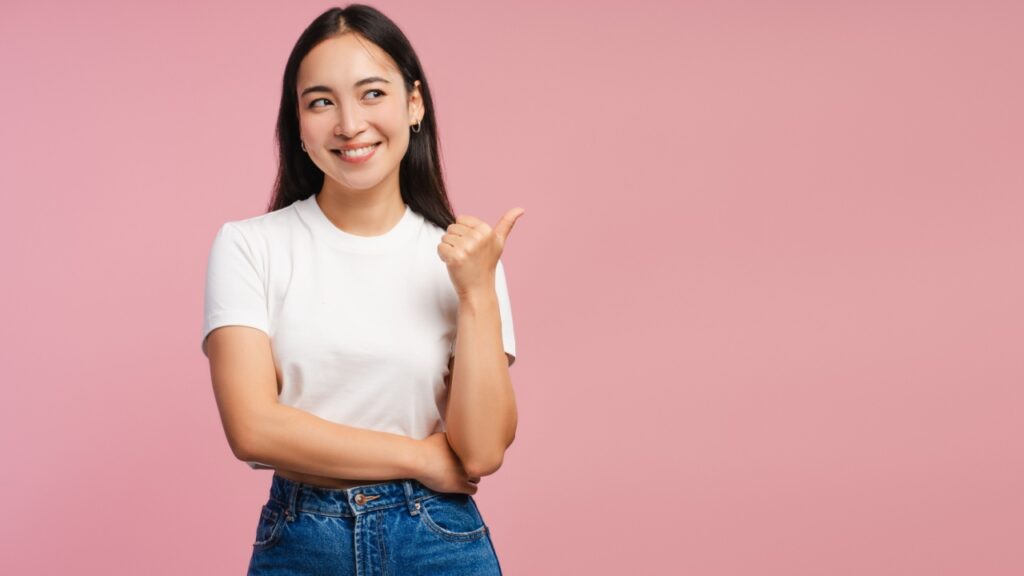 mulher sorrindo e com o polegar direito apontado para o lado em fundo rosé