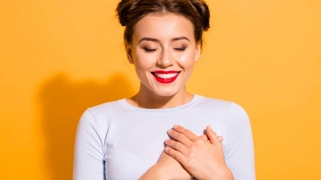 Retrato de uma mulher alegre, com as mãos tocando o coração, isolada sobre um fundo amarelo.