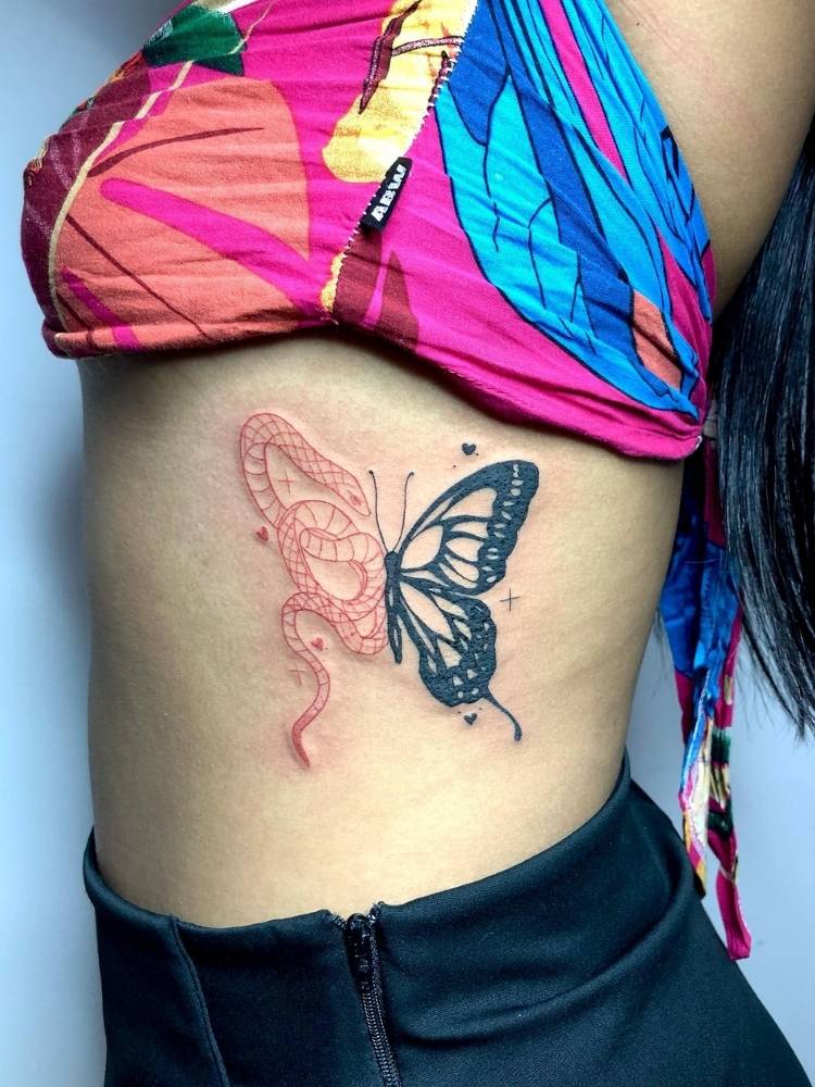 borboleta-costela-cobra Tatuagem de uma asa de borboleta com traços grossos e pretos e a outra asa é o desenho de uma cobra de traços finos na cor vermelha. A tattoo foi feita na costela de uma mulher de pele clara, usando uma saia preta e uma blusa rosa, laranja e azul.