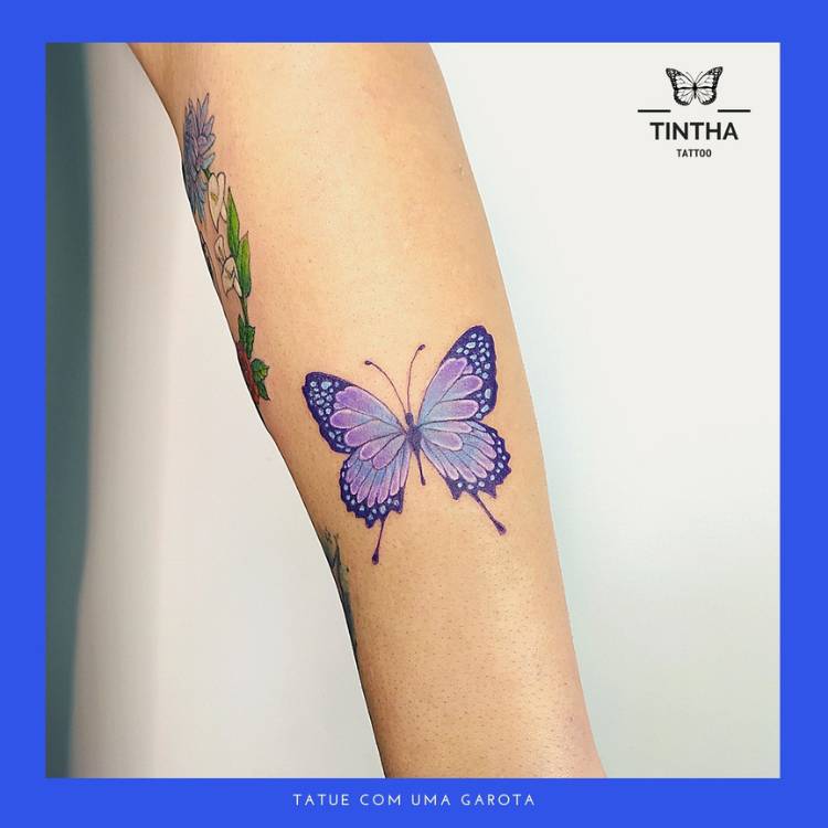 colorida-azul-roxo-braco Tatuagem de borboleta em traços finos com tons de azul e roxo no braço de uma mulher de pele clara.