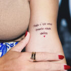 destaque-frases-para-tatuagem-140x140-1-2 A imagem apresenta uma tatuagem delicada no braço de uma mulher com a frase: "Nada é tão nosso quanto nossos sonhos". Abaixo da frase, há um pequeno coração vermelho. Detalhes incluem unhas pintadas e acessórios elegantes.