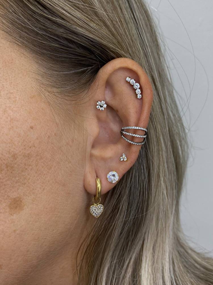 Orelha com piercing de flor no anti hélix, perfuração no helix, conch, flor abaixo do conch e dois furos no lóbulo, sendo o primeiro uma argola com coração cravejado.