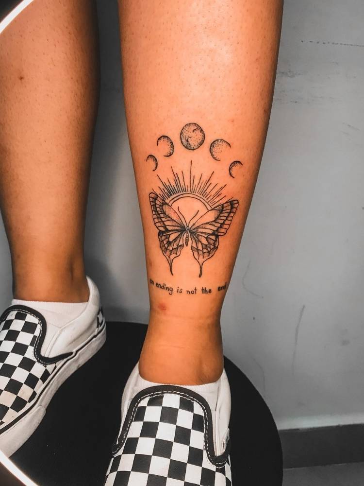 frase-borboleta0perna Tatuagem de Borboleta cheia de detalhes, sombreamento, traços finos e mais grossos. Ela acompanha a frase "An ending is not the end" e as fases da lua acima. Foi feita na canela de uma mulher de pele clara que está usando um tênis quadriculado de branco e preto.