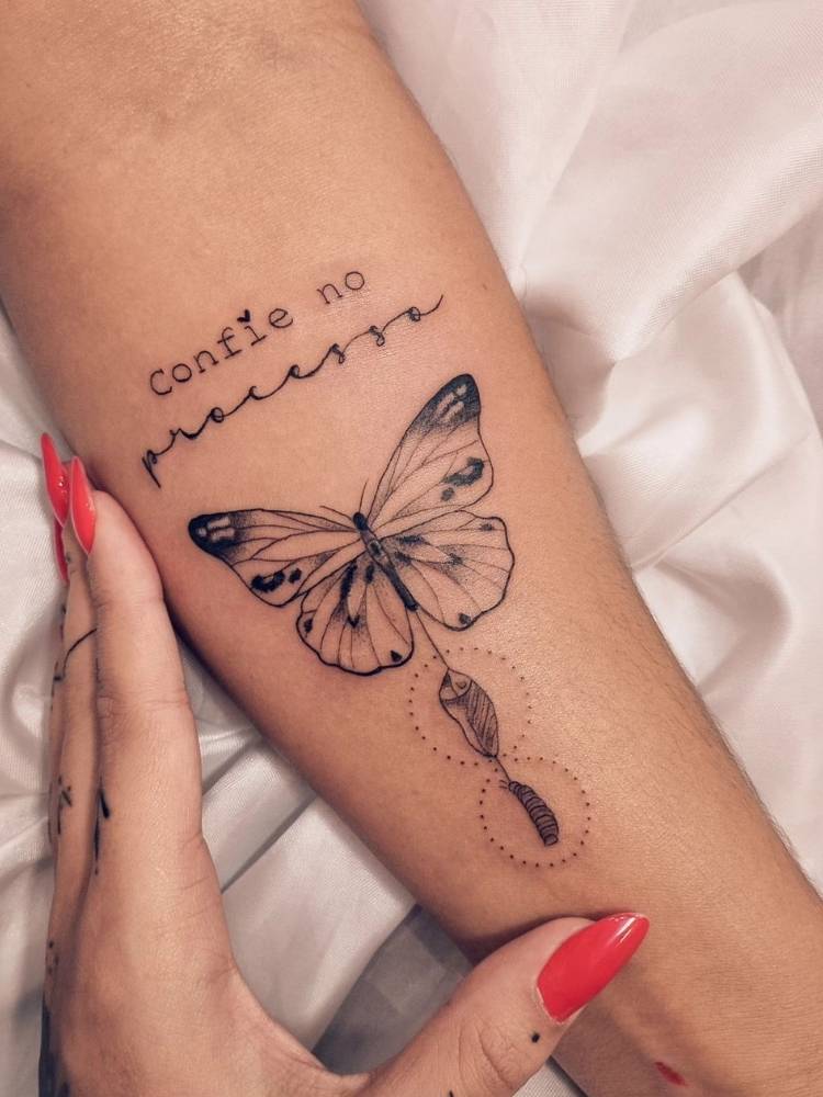 frase-com-borboleta-metamorfose Tatuagem de borboleta com sombreamento, traços finos e grossos. Nela, há o desenho do casulo de onde e a borboleta saiu e também a frase "Confie no processo". Foi feita no braço de uma mulher de pele morena.