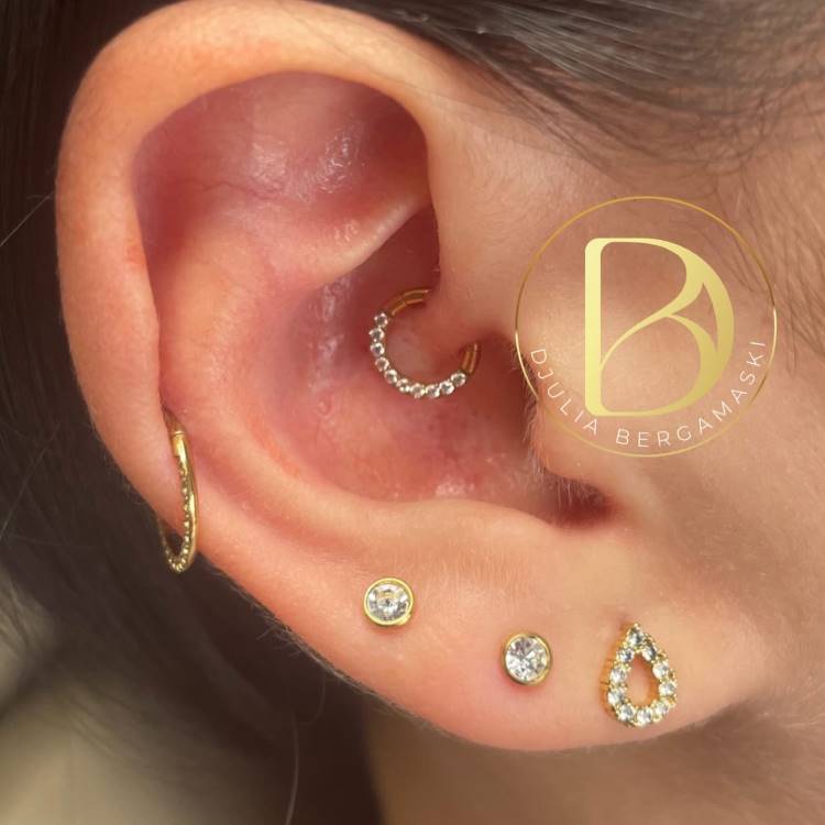 Orelha com piercing em dourado no hélix, com argola cravejada no daith e pequenos brincos no lóbulo.