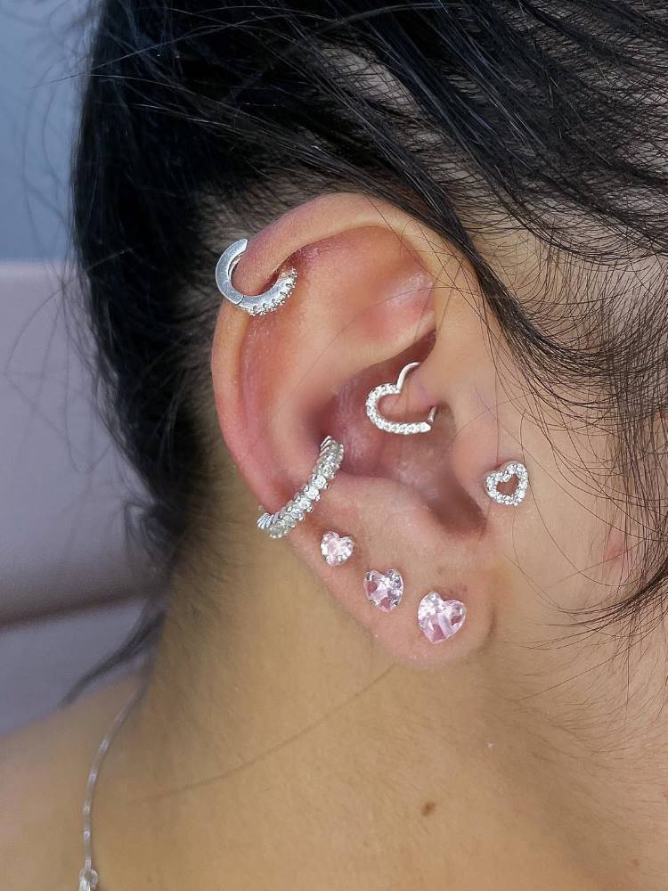 Orelha com piercing na hélix, argola de coração no daith, coração no tragus, argola no conch e três brincos pequenos no lóbulo.
