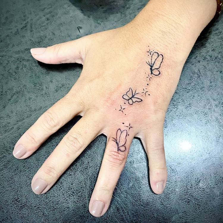 mao-delicada-borboleta Tatuagem de várias borboletas pequenas no dorso e dedo anelar da mão de uma mulher de pele clara.