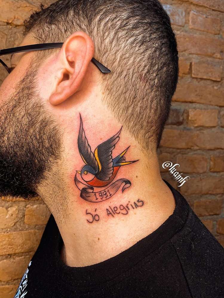 Homem de pele clara com andorinha colorida old school no pescoço com faixa escrito "1991" e "só alegrias"