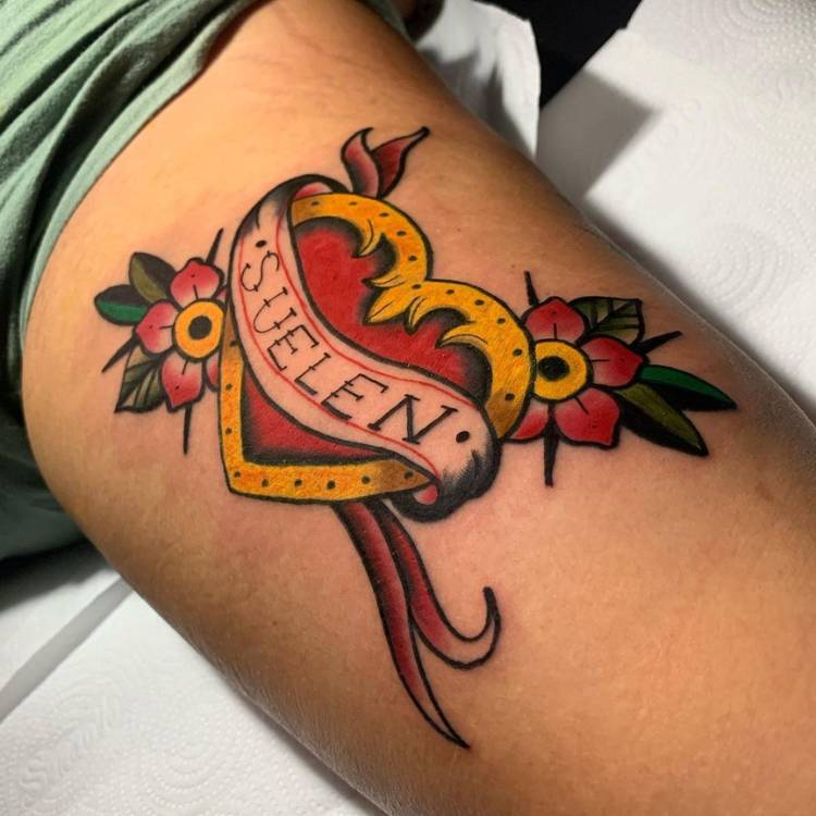 Homem de pele clara com tatuagem old school de coração vermelho com flores escrito "Suellen"