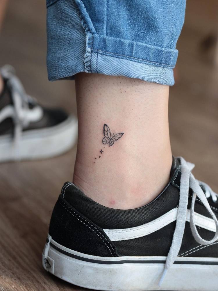 pequena-canella-tattoo-borboleta Tatuagem pequena de uma borboleta em traços finos no tornozelo de uma mulher de pele clara usando calça jeans e tênis preto.