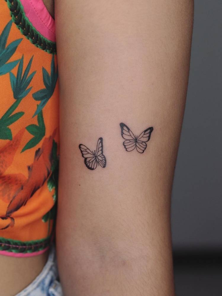pequena-tattoo-braco-borboleta Tatuagem de duas borboletas pequenas, delicadas com traço fino no interior do braço de uma mulher de pele clara e vestindo uma blusa laranja e verde.