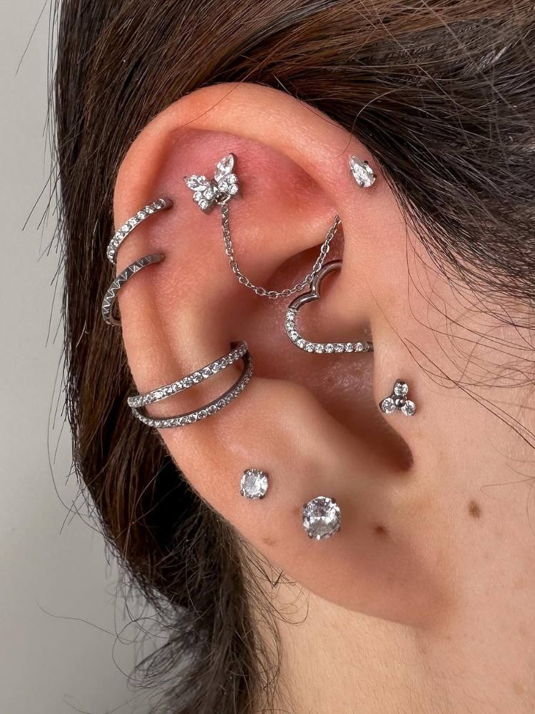 Orelha de pele clara com piercing de borboleta na scapha com corrente até ponto de luz no anti helix, duplo helix com argolas, argola dupla no conch, dois pontos de luz no lóbulo e um no tragus