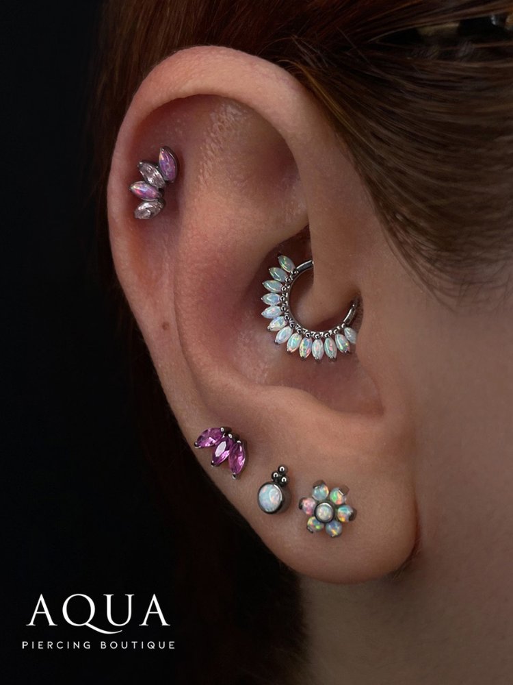 Orelha de pele clara com joias florais, sendo meia flor lilás no hélix, argola de pétalas opala no daith e lóbulo com piercing 3 pétalas liás, bola opada e flor