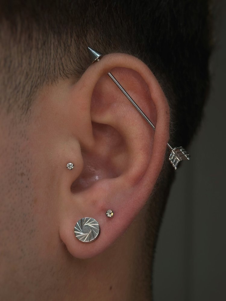 Orelha de pele clara com piercing transversal de flecha, ponto de luz no tragus, brinco prata no lóbulo e ponto de luz no segundo furo