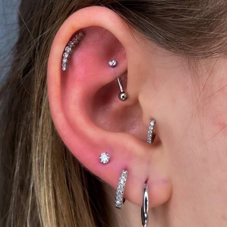 Orelha com piercing na hélix, rook, tragus e três brincos no lóbulo,sendo duas argolas e um ponto de luz, tudo no prateado.