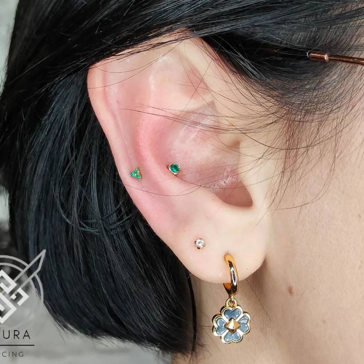 Orelha com piercing com pedra verde no snug e dois brincos no lóbulo, sendo um ponto de luz e outro uma argola dourada ocm flor azul
