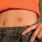 tattoo-cintura-140x140-1-2 A imagem mostra a parte inferior do abdômen de uma pessoa, com um top laranja e jeans. Há um piercing no umbigo e uma tatuagem delicada de flor com traços finos e pétalas vermelhas. A mão está levemente posicionada sobre o cós da calça, destacando o estilo casual e jovem.