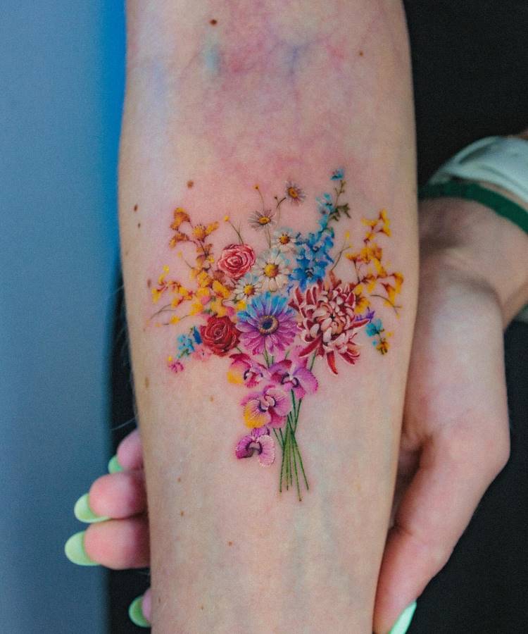 Tatuagem colorida no antebraço com um buquê de flores detalhado. Inclui rosas, margaridas, crisântemos e flores lilases, em tons de vermelho, azul, rosa, amarelo e roxo. Os detalhes são delicados, com hastes verdes. Fundo neutro e mão visível.