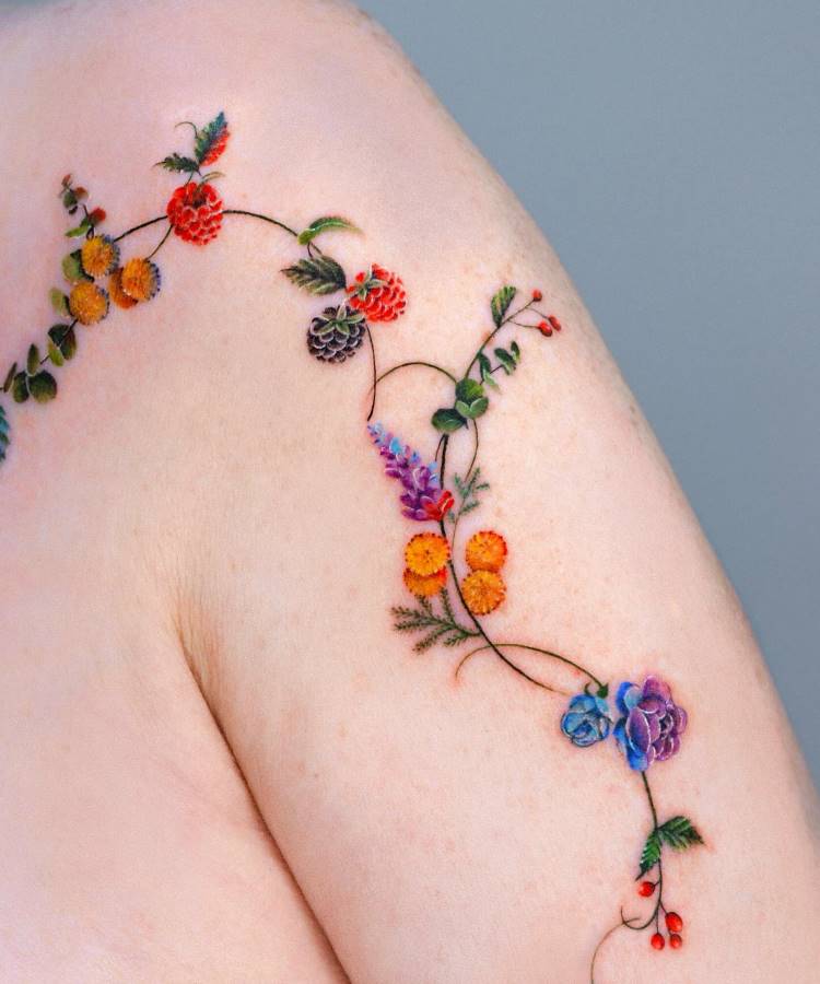 Tatuagem delicada no ombro, com flores coloridas, como rosas, lavandas e margaridas em tons vibrantes de azul, roxo, laranja e vermelho. Ramos e folhas verdes conectam os elementos, desenhando um padrão orgânico e detalhado. Fundo cinza neutro.