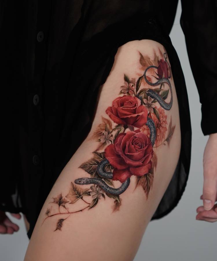 Tatuagem detalhada na coxa com duas rosas vermelhas vibrantes rodeadas por folhas em tons outonais. Uma serpente azul-acinzentada entrelaça-se pelas flores. A pele está parcialmente coberta por uma roupa preta translúcida, destacando o trabalho artístico.