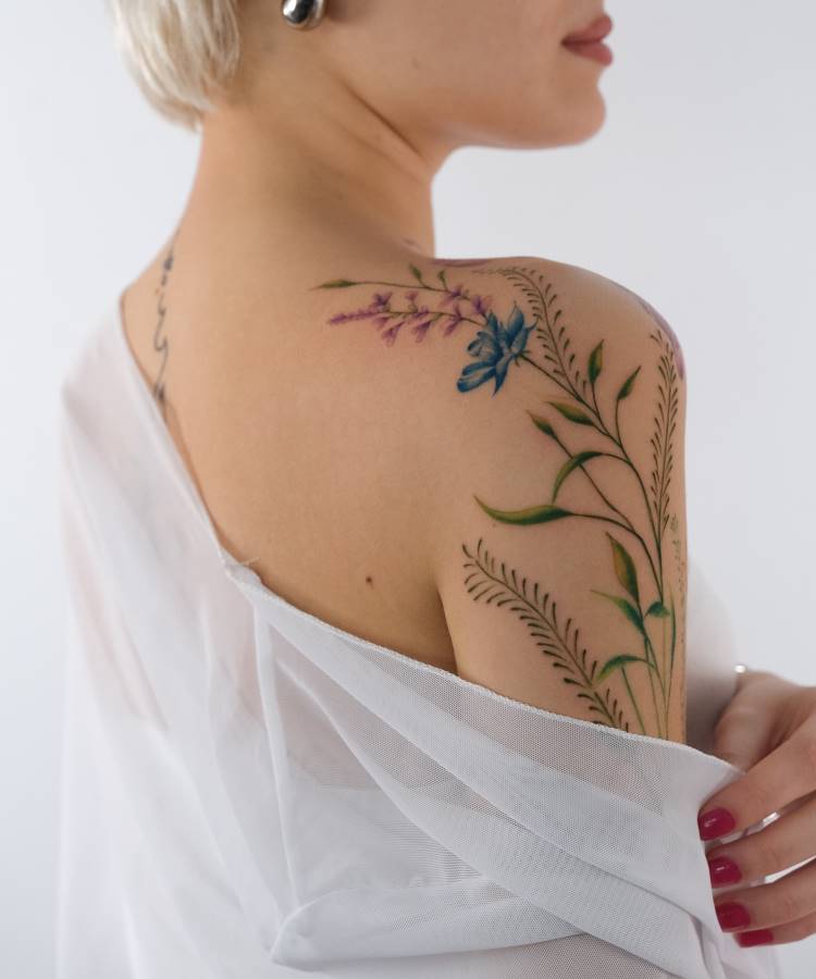 Tatuagem delicada no ombro com flores coloridas, incluindo uma azul e outra lilás, cercadas por folhas verdes detalhadas. A pessoa usa tecido translúcido branco, revelando parcialmente a tatuagem. Fundo branco e cabelo curto completam a composição elegante.