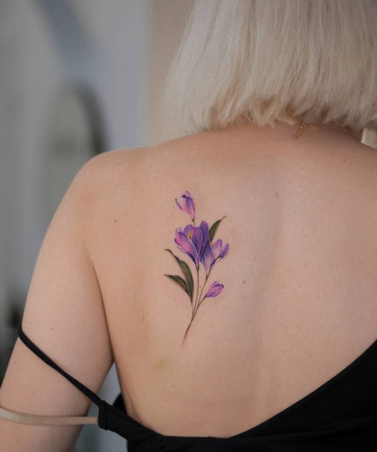 Tatuagem delicada nas costas com flores de crocus em tons vibrantes de roxo, acompanhadas por folhas verdes. Traços finos e realistas destacam a elegância do desenho. A pessoa de cabelos loiros usa uma blusa preta com alças finas.