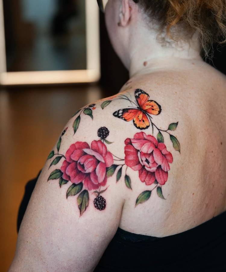 Tatuagem colorida no ombro com flores rosas vibrantes, folhas verdes e pequenos frutos escuros. Uma borboleta laranja com detalhes pretos está posicionada acima, criando equilíbrio. Fundo desfocado com reflexo de espelho e tons quentes ao redor.