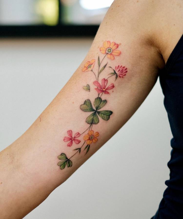 Tatuagem delicada no braço com flores coloridas em tons de amarelo, rosa e vermelho, acompanhadas por folhas verdes e trevos de quatro folhas. Os traços são finos e realistas. Fundo neutro e pessoa vestindo uma regata preta.