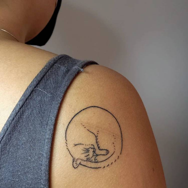 tatuagem-feminina-ombro-atras-gato Mulher de pele morena com regata cinza e tatuagem na parte de trás do ombro, com o desenho redondo de um gato se lambendo
