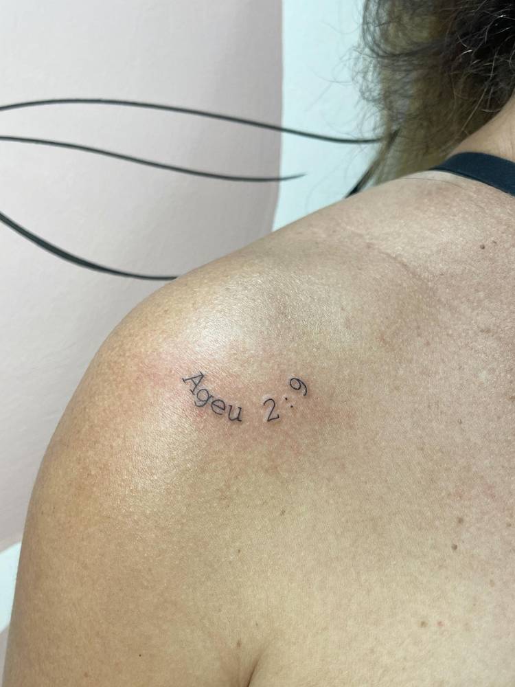tatuagem-feminina-ombro-delicada-bibilia Mulher de pele clara com tatuagem escrita fine line "Ageu 2:9"