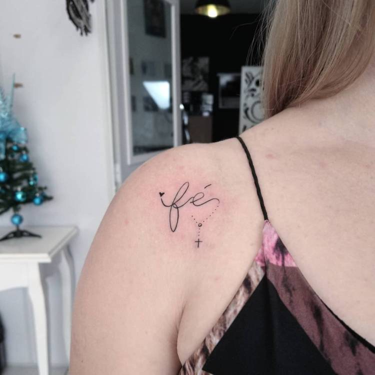 tatuagem-feminina-ombro-delicada-fe Mulher loira de pele clara usando regata preta e com tatuagem fine line escrito "fé"