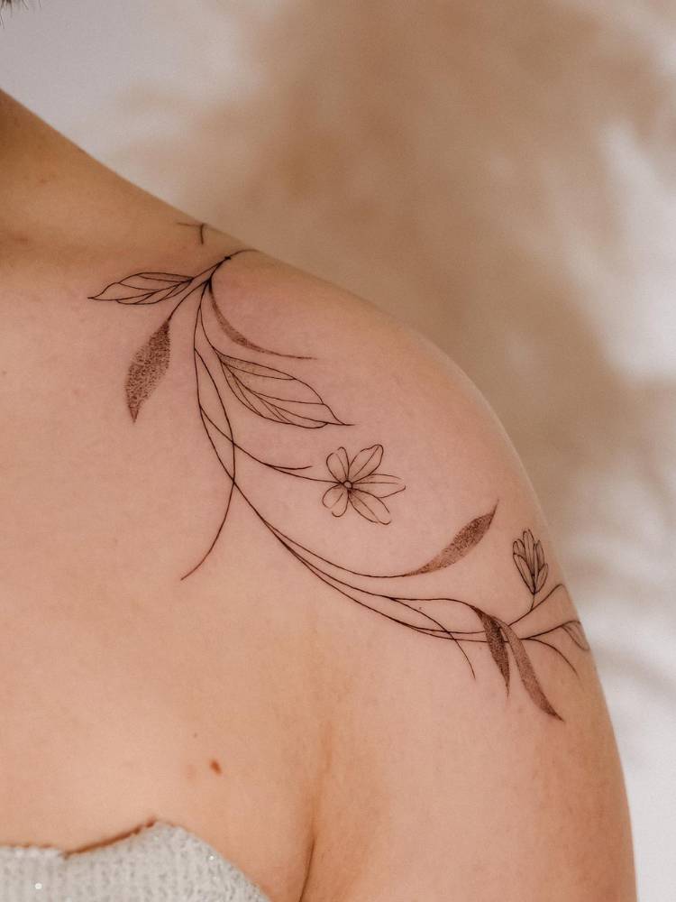 tatuagem-feminina-ombro-delicada-flores Mulher de pele clara com ombro tatuado com ramos de flores ao redor dessa parte do corpo