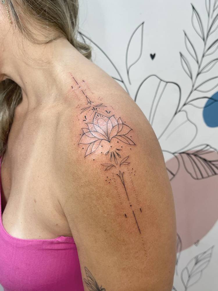 tatuagem-feminina-ombro-delicada-lirio Mulher de pele clara de lado, usando blusa rosa e com tatuagem de lírio no ombro, com linhas e ornamentos