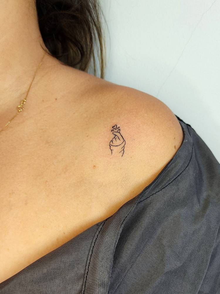 tatuagem-feminina-ombro-delicada-mao Mulher de pele clara e blusa preta com tatuagem de mão fazendo coração coreano no ombro