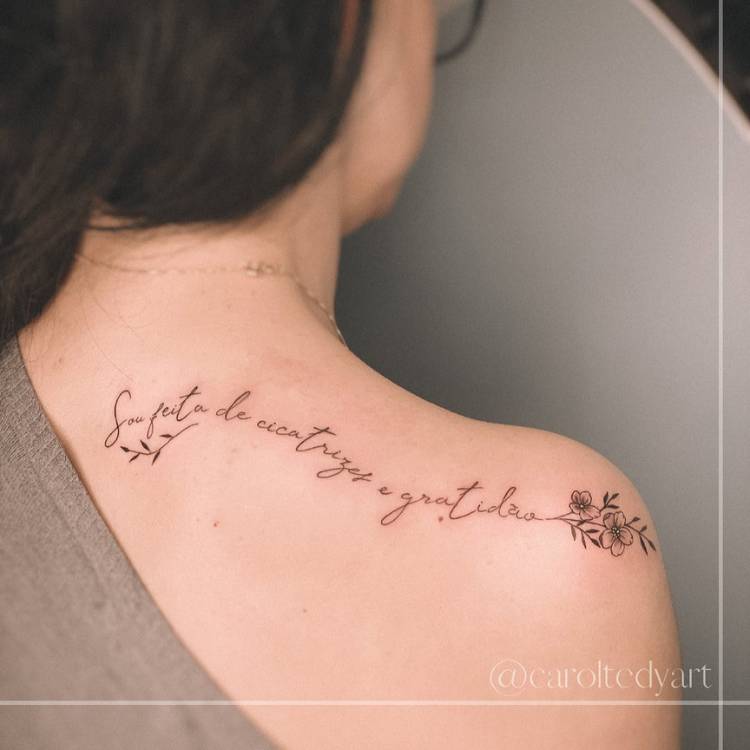 tatuagem-feminina-ombro-frase-atras Mulher de pele clara de costas usando blusa cinza e tatuagem "Sou feita de cicatrizes e gratidão" com flor