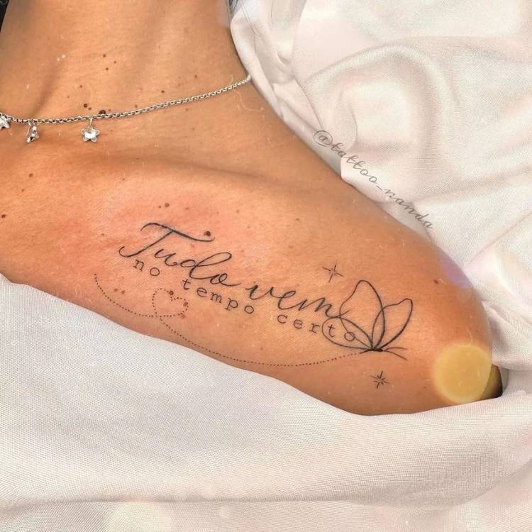 tatuagem-feminina-ombro-frase-tudo-vem Mulher de pele clara com corrente prateada e tatuagem de frase escrito "tudo vem no tempo certo" com borboleta.
