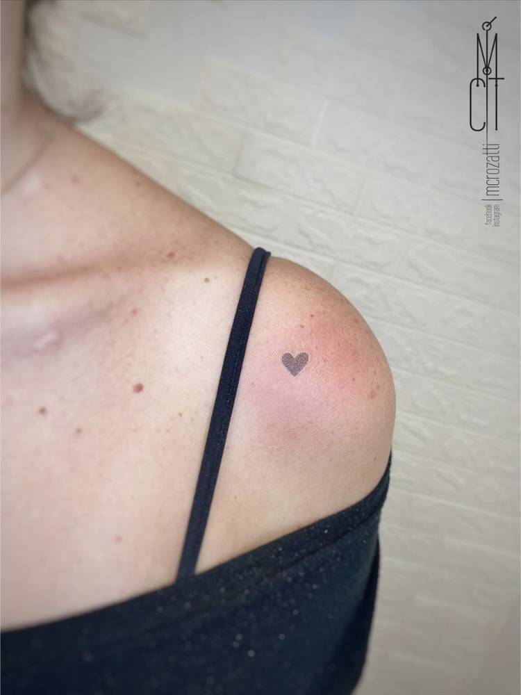 tatuagem-feminina-ombro-mini-coracao Mulher de blusa preta no ombro e sutiã preto com tatuagem de mini coração sombreado no ombro