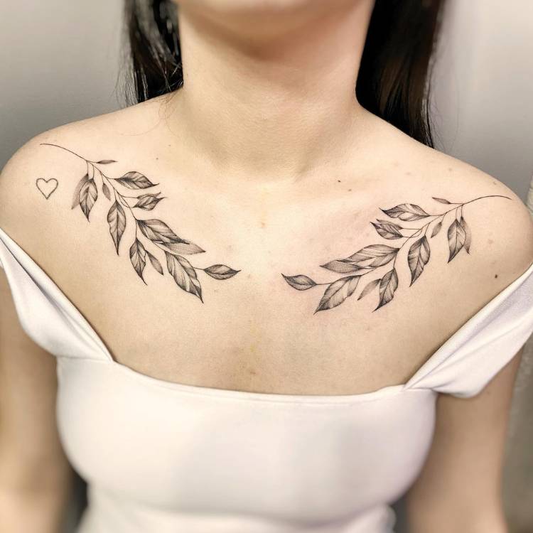 tatuagem-feminina-ombro-ramos Mulher de pele clara e cabelo escuro usando regata bege e tendo dois ramos tatuados, um em cada ombro, além de um coração embaixo de um dos ramos