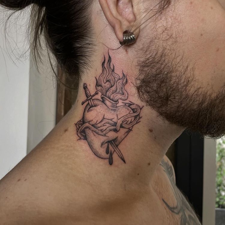 tatuagem-masculina-pescoco-10-1 A imagem mostra uma pessoa de perfil com cabelo preso e barba. Ela tem uma tatuagem no pescoço de um coração anatômico com chamas saindo do topo e uma espada atravessando-o. A tatuagem é detalhada em preto e cobre a lateral do pescoço até a parte de trás da orelha.