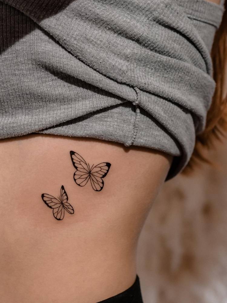 traco-fino-borbolet Tatuagem de duas borboletas em traço fino com um leve traço grosso na costela de uma mulher de pele clara usando uma blusa cinza.