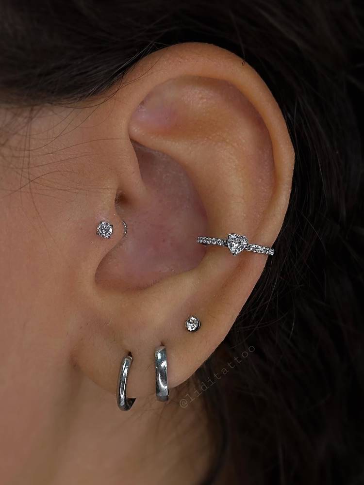 Orelha com piercing no tragus, conch com coração e três furos no lóbulo, todos em prata, sendo os dois primeiros argolas e o terceiro um brilho.